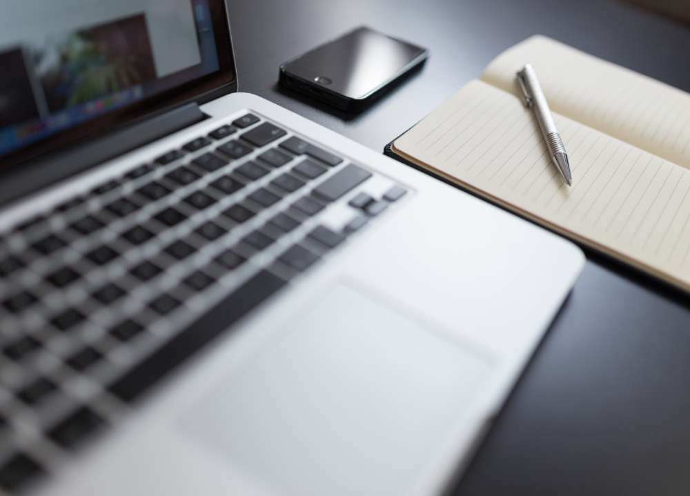 Laptop sitting on desk with notepad, pen, and iPhone.